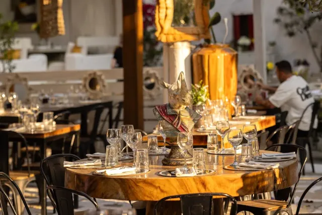 A round polished wooden table with wine glasses and a ceramic figurine, and metal chairs in warm light