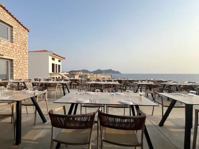 Tables set on a paved terrace facing the sea, with a headland and a stone structure on the horizon