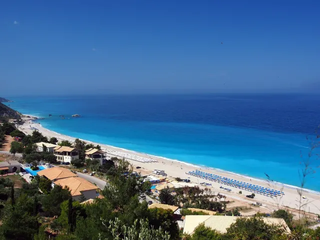 A long sandy beach with water in shades of blue, houses on the green slope above
