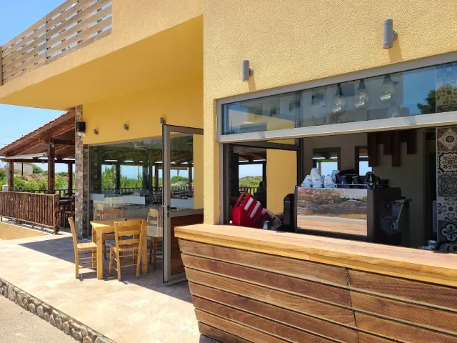 A yellow building with a large glass window, a wooden counter and a pergola with tables beside it
