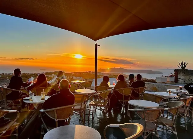 People seated around tables under a dark canopy, facing a setting sun above the coastline