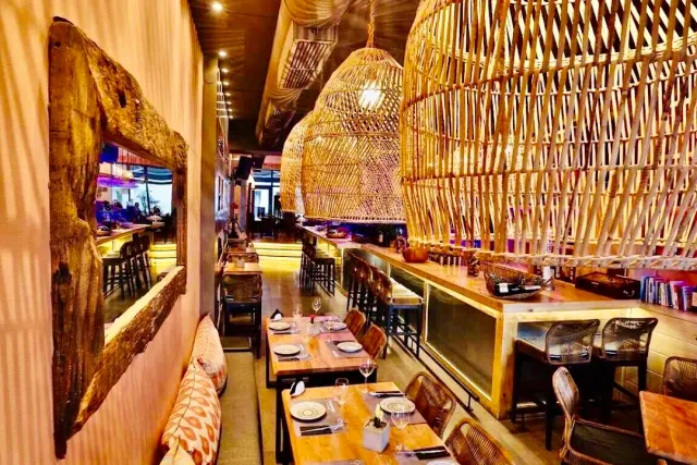 An interior with large wicker pendant lamps, laid wooden tables and a lit bar counter