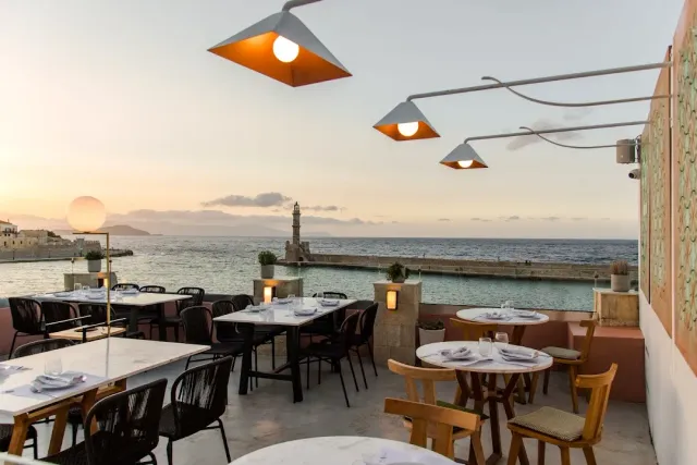 A terrace with hanging lamps and marble tables, and a lighthouse on a pier at sunset