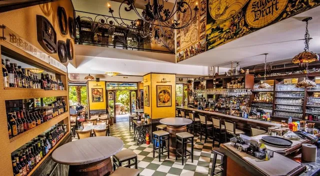 An ornate bar with a checkered floor, shelves of bottles, chandeliers and an upper floor gallery
