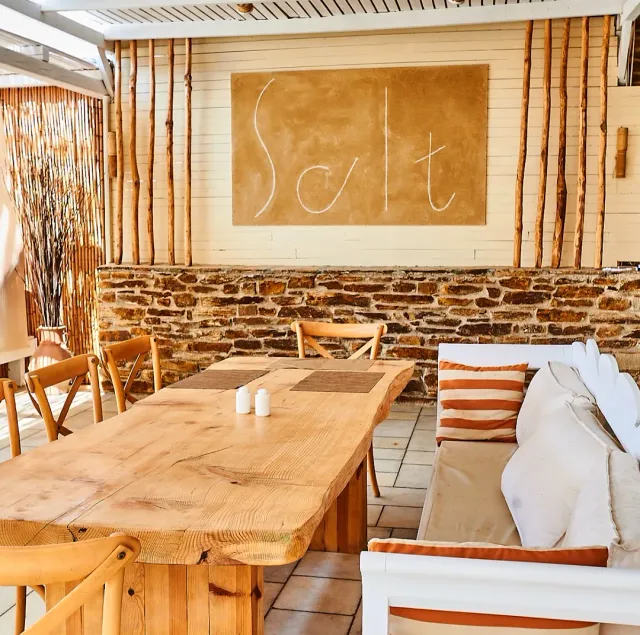 A long wooden table and chairs facing a stone wall, a bench with orange striped cushions and bamboo poles