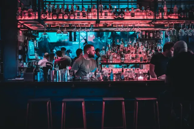 A bar counter in pink and blue neon lighting, bartenders and bottle shelves behind