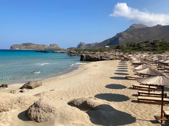 A row of straw umbrellas and wooden loungers on wide sand, beside a turquoise sea and mountains