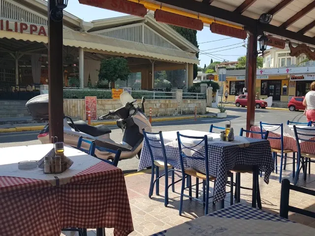 A covered veranda with checked tablecloths and blue chairs, and a scooter parked on the street