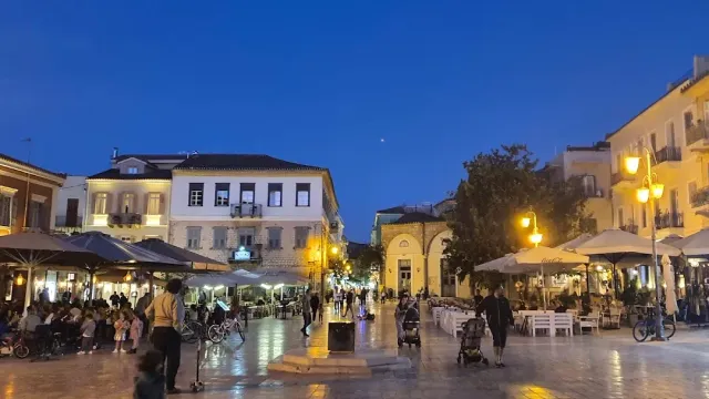 A paved square in the evening, lit lamps, cafe umbrellas and illuminated buildings