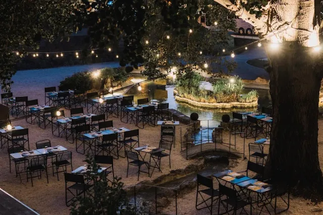 Black metal tables and chairs among trees, with a string of lights and a pond