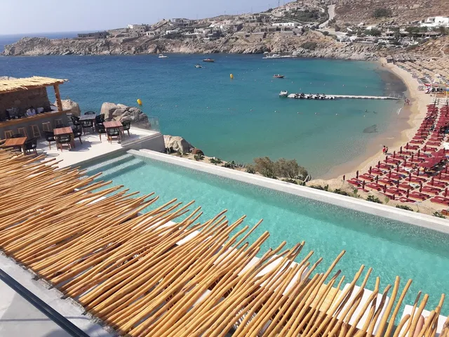 An infinity pool above a turquoise bay, with rows of red sun loungers on the beach