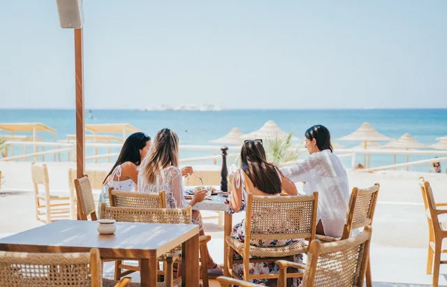 Four women sit in wicker chairs at a table, facing straw parasols and the sea
