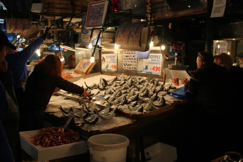 A lit fish stall in a covered market, piles of fresh fish and price signs in Greek