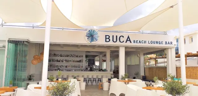 An open frontage under white shade sails, a counter with bottle shelves and white armchairs