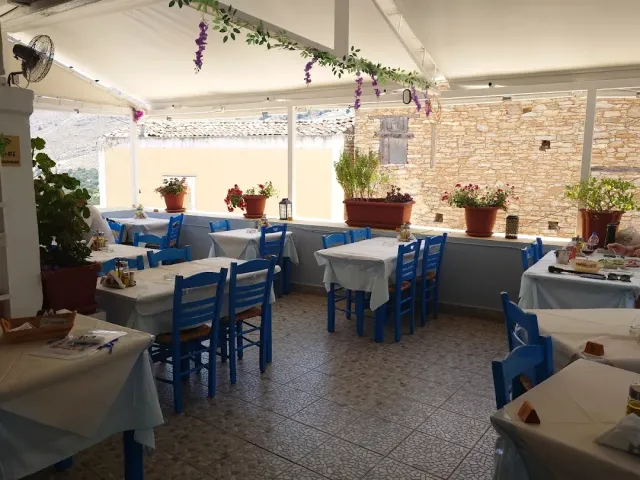 A covered terrace with blue chairs, white cloths and flowering geranium pots