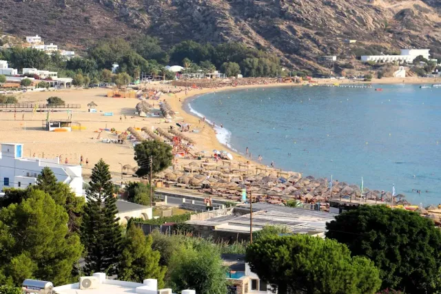 An elevated view of a curving sandy bay with rows of straw umbrellas and white buildings