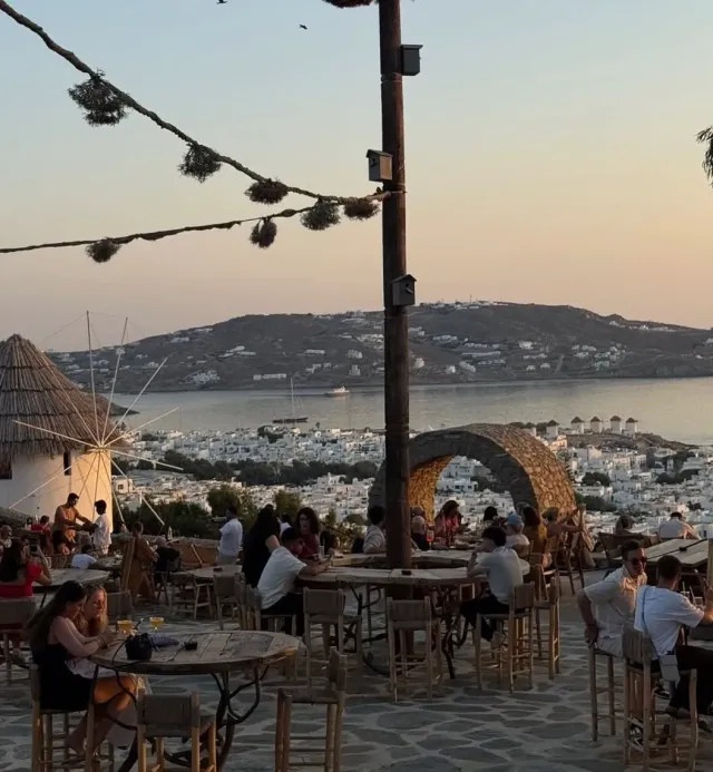 People seated on a paved terrace at sunset, facing a white town, a bay and a windmill