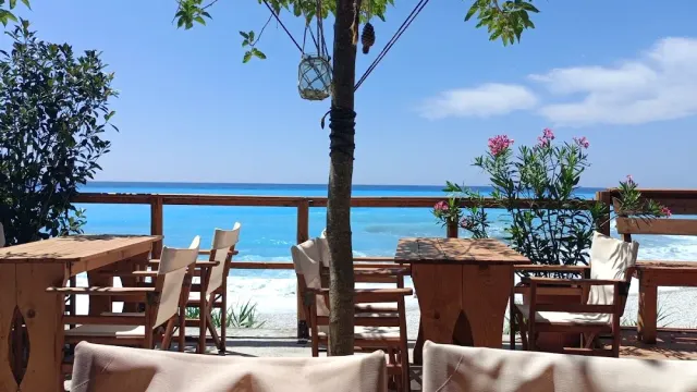 Wooden tables on a deck facing a white beach and a turquoise sea, with pink blossom