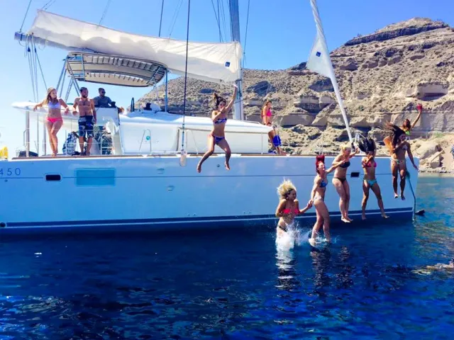 A white catamaran with passengers jumping from the deck into blue water, a cliff behind