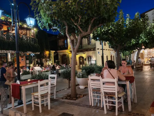 White tables in a paved square in the evening, under trees and lit street lamps