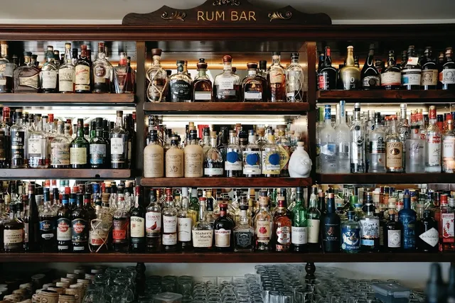 Wooden shelves packed with liquor bottles above rows of glasses, beneath a carved sign