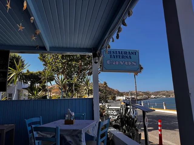 A table with blue chairs under a wooden roof, facing a taverna sign, a coastal road and the sea