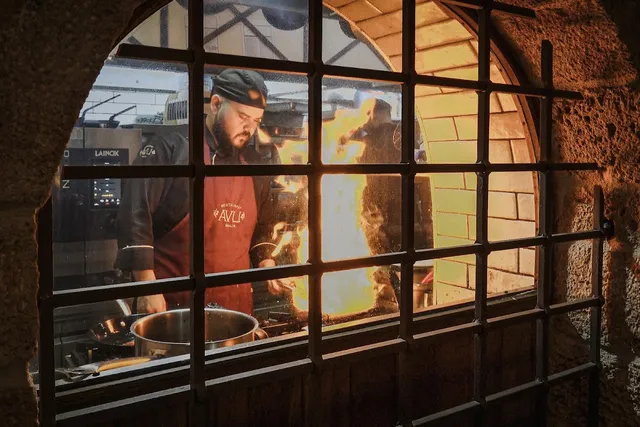 A cook in a red apron cooking over flames, seen through a barred window in a stone wall