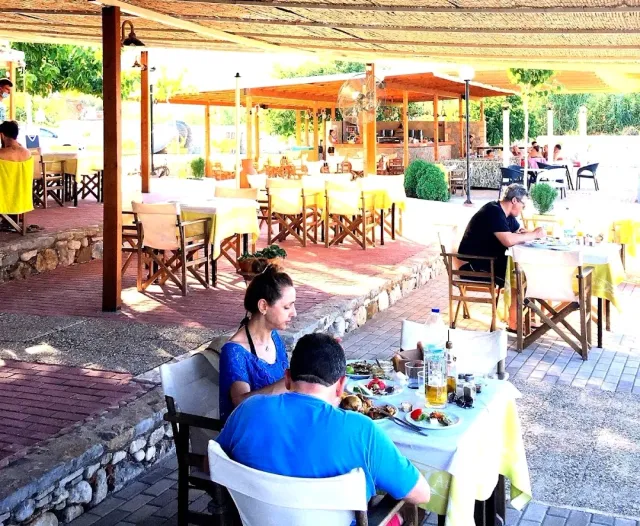 A shaded dining area under a reed canopy with yellow tablecloths, wooden chairs and diners at a meal