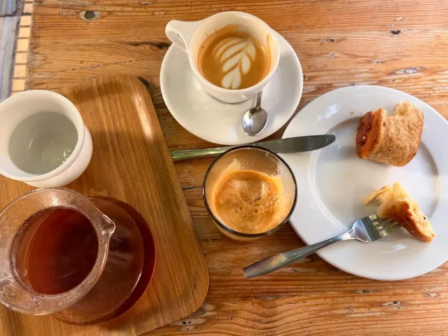 A view from above of a wooden table with a cappuccino, an espresso, a glass of tea and a pastry