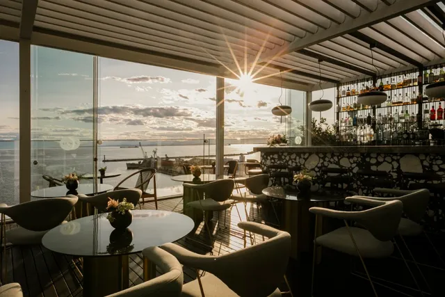 A covered terrace with round tables and glass walls facing the bay at sunset