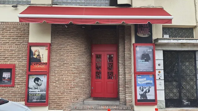 A red entrance door in a brick wall, with a red awning and concert posters on either side