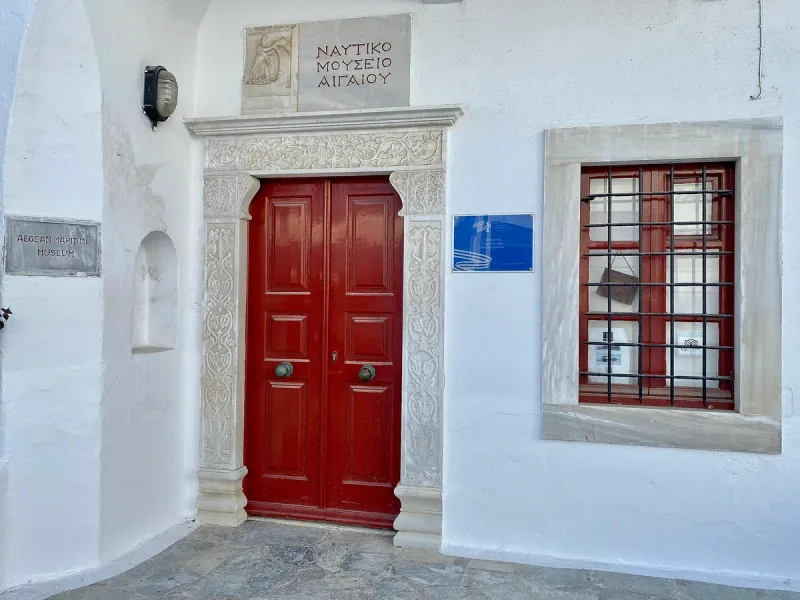 The white facade of a Cycladic house with a red wooden door and a carved Greek inscription