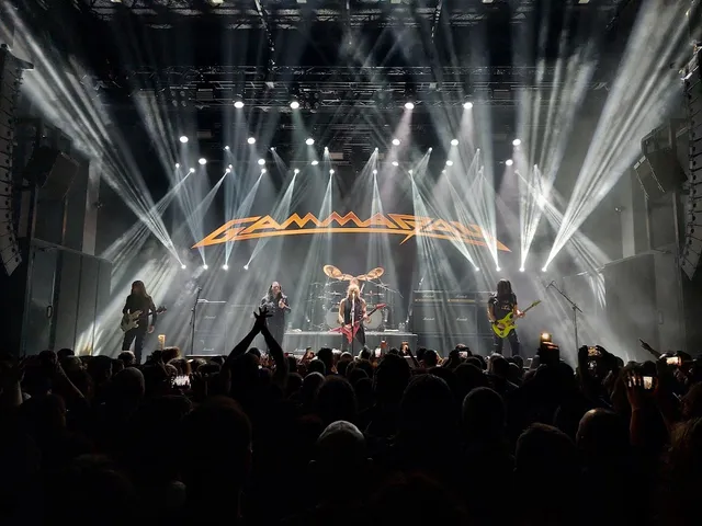 A band performs on a large stage in white light beams, before a dense crowd in the dark