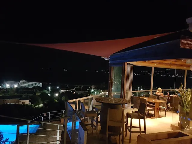 A lit rooftop terrace at night with high tables, above a pool and the lights of the coast