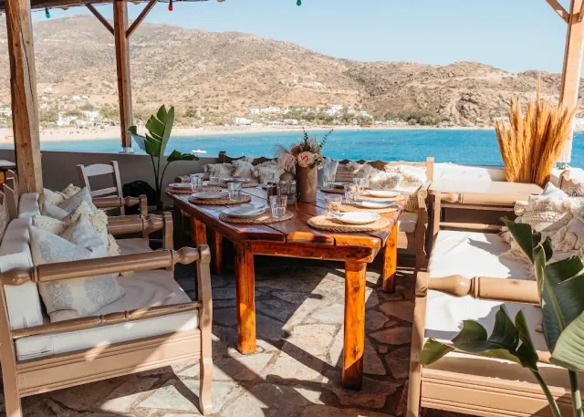 A long laid wooden table on a terrace with white armchairs, facing a sandy bay and hills
