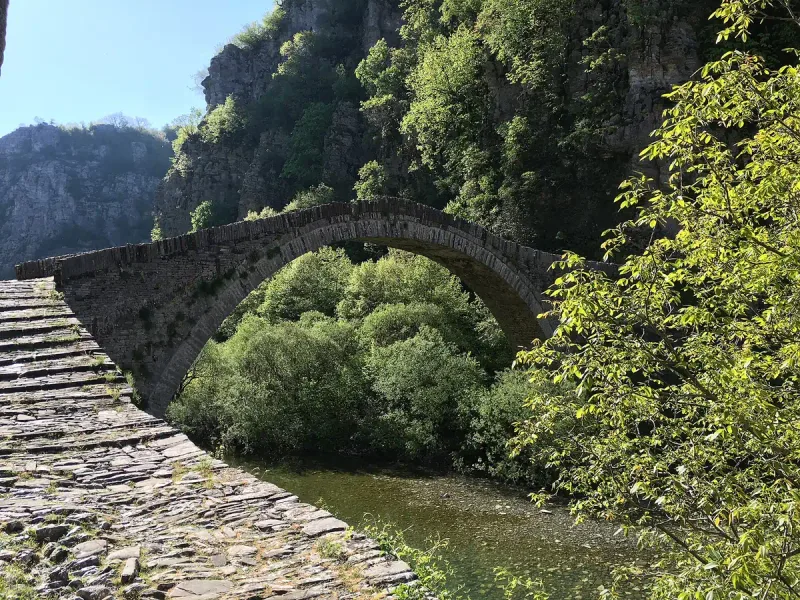 A single arch stone bridge over a stream, with a paved walkway and dense greenery around it