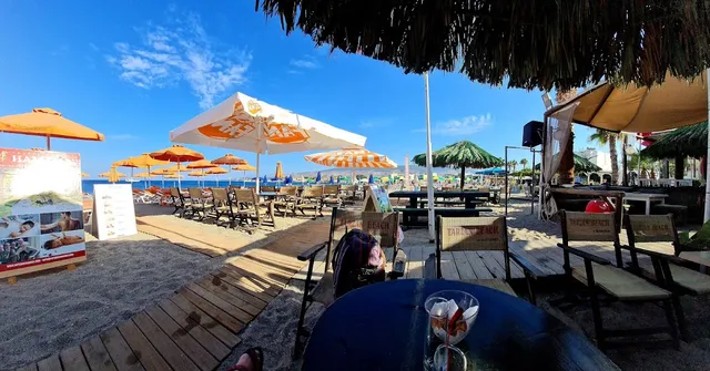 Wooden tables and chairs on a boardwalk on a sandy beach, with orange umbrellas and blue skies