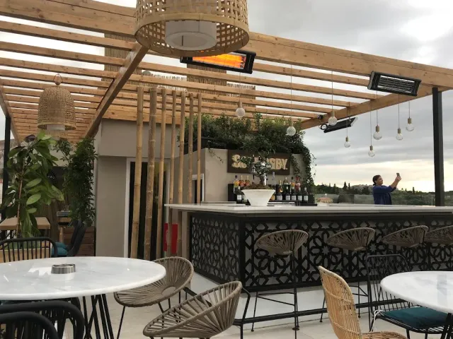 A rooftop bar with a wooden pergola, hanging straw lamps and tall stools beside the counter