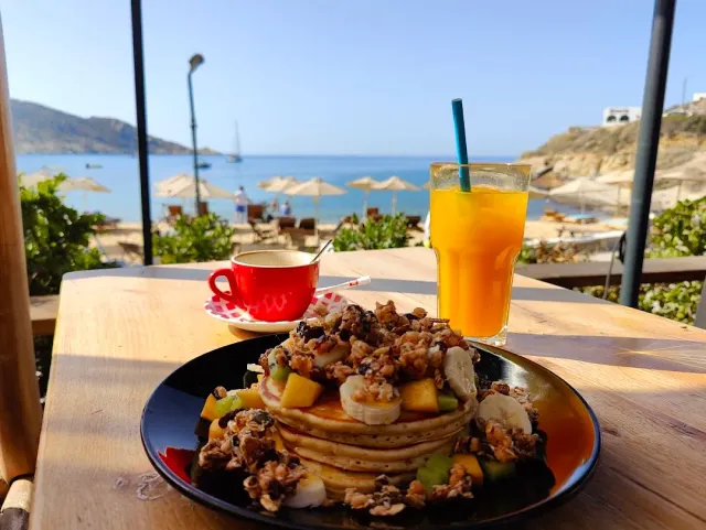 A stack of pancakes with granola and fruit, a red cup and juice, and a shaded beach behind