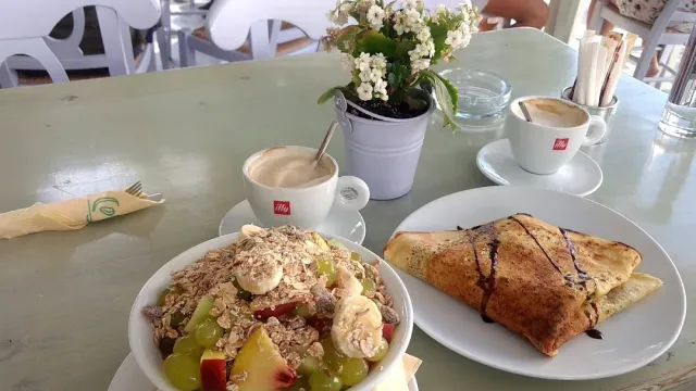 A bowl of fruit with granola, a crepe with chocolate sauce and two coffee cups on a green table