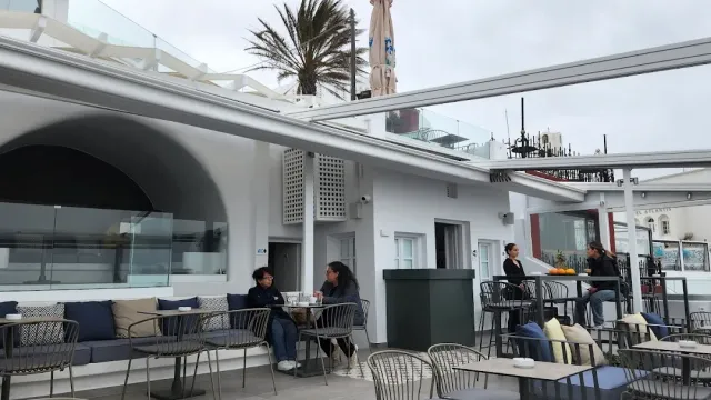 A white terrace with a retractable shade, sofas, round tables and a palm behind