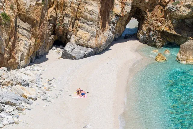 A white pebble beach between cliffs, with a natural rock arch dividing two coves