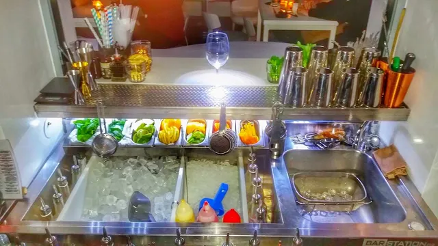 A stainless steel bar station with ice, cut fruit and shakers lined up in a row