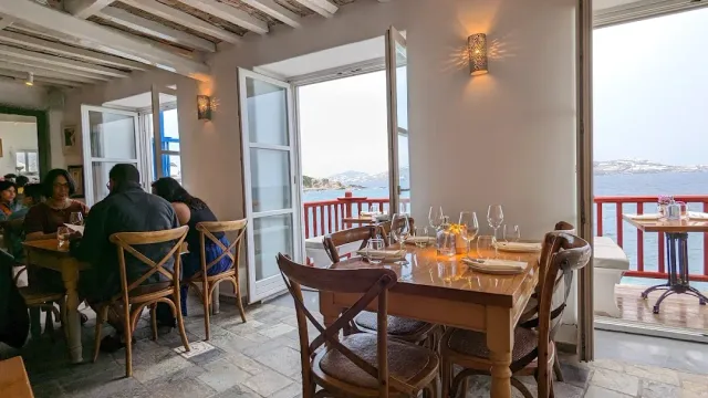 A room with set wooden tables and a door open onto a balcony with a red railing facing the sea