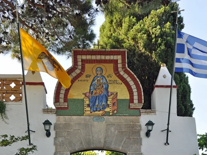 A whitewashed monastery gateway with a mosaic icon and flags flying beside it