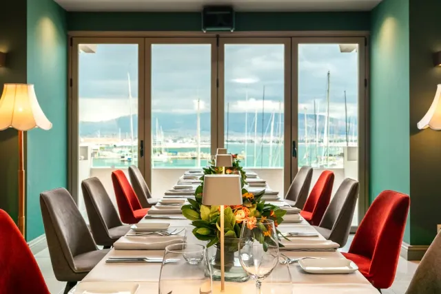 A long set table in a green room with red chairs and a window onto a marina