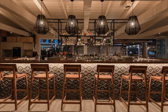 A long bar counter with wooden stools, hanging woven lamps and shelves of bottles