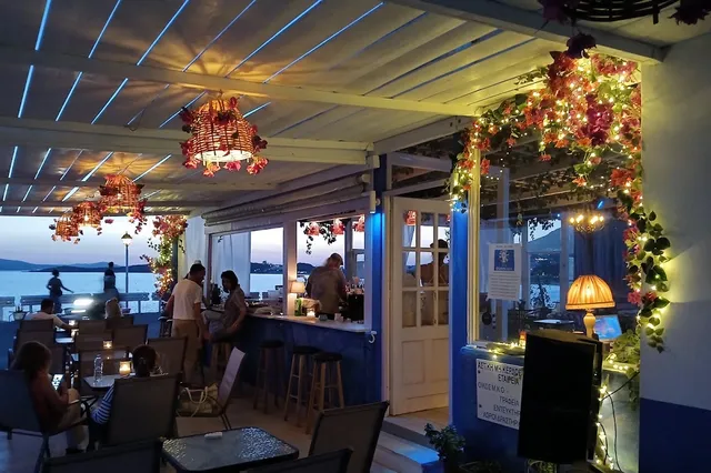 A blue and white bar with flower garlands and hanging lamps, and a bay in the background at sunset