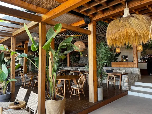A wooden pergola with a large straw lampshade, banana plants, round tables and a stone counter