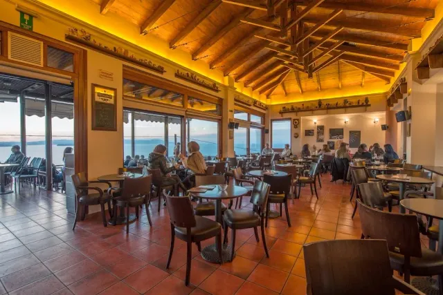 A wide hall with a wooden beam ceiling, yellow lighting, tables and a sea view in the windows